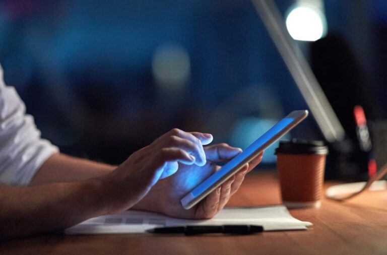 Close‑up of hands using a tablet at a desk in a dimly lit workspace, representing a career journey into cybersecurity with Ergo.